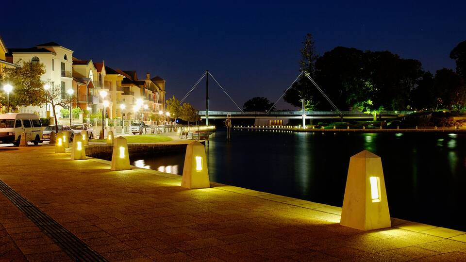A nighttime riverside walkway with illuminated bollards, a pedestrian bridge, and buildings along the riverbank.