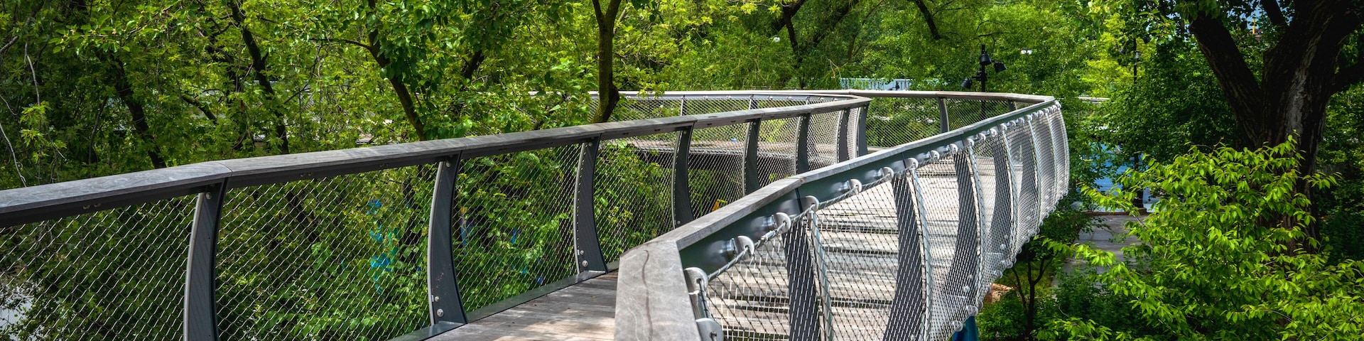 A wooden pedestrian bridge with metal railings, surrounded by lush green trees.