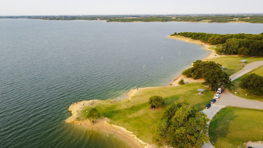 Un paysage côtier comprenant une étendue d’eau importante, une plage de sable et un espace de stationnement avec des véhicules.