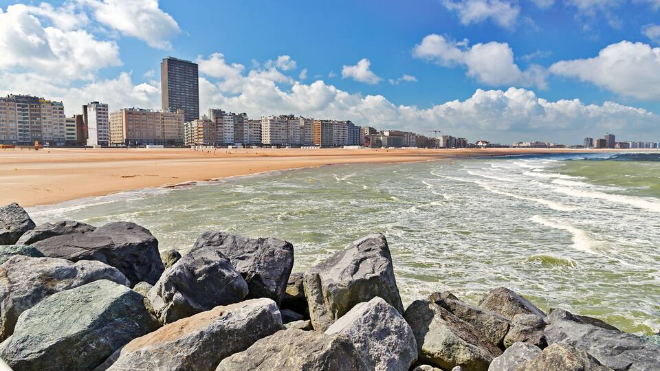 A beach with large rocks in the foreground, a sandy shore, and a city skyline in the background.