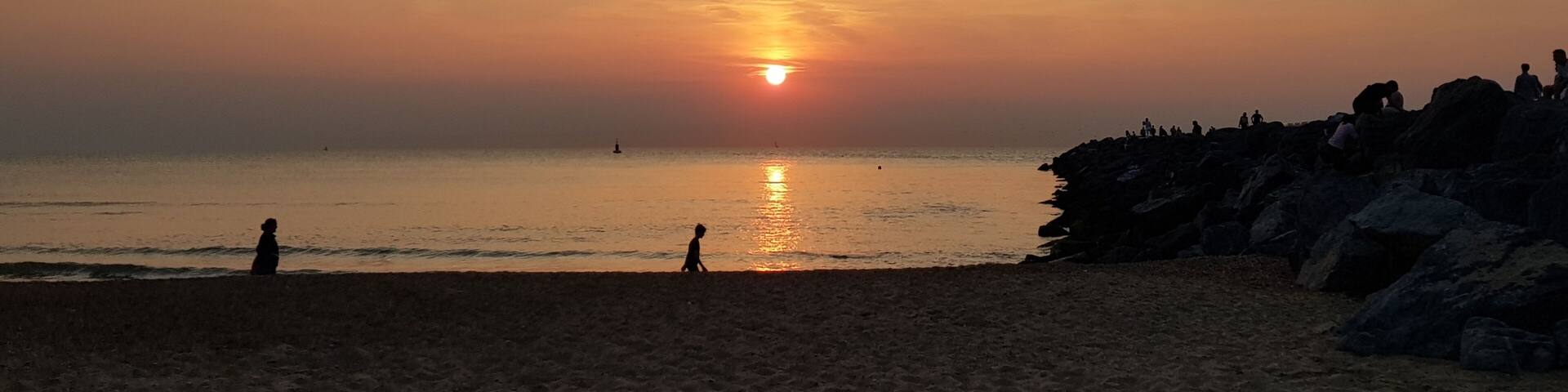 A beach at sunset with the sun near the horizon, silhouettes of people, and a rocky outcrop on the right.