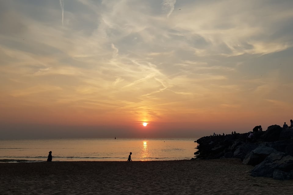 A beach at sunset with the sun near the horizon, silhouettes of people, and a rocky outcrop on the right.