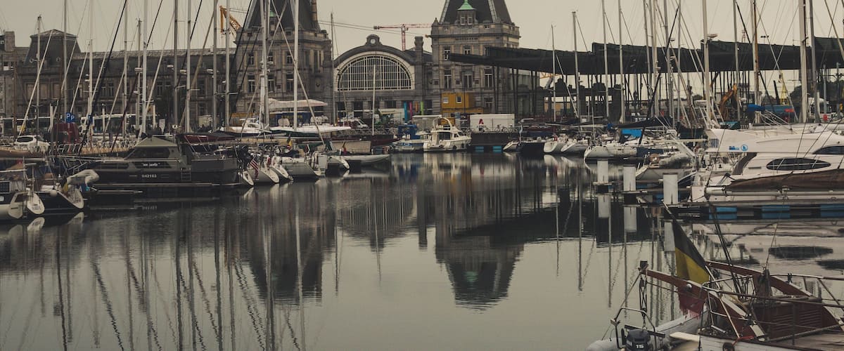 A marina with numerous boats docked, a large building with a distinctive dome, and calm water reflecting the boats.