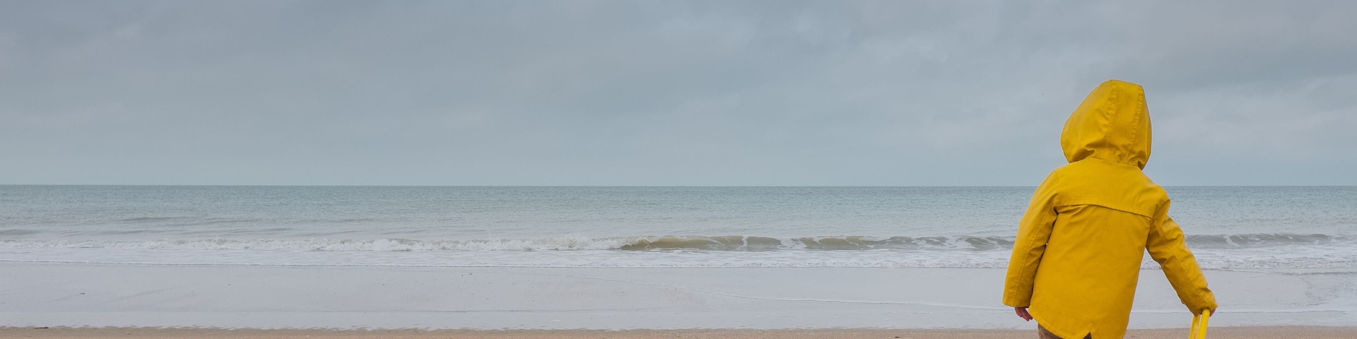 A person in a yellow raincoat standing on a sandy beach facing the ocean.