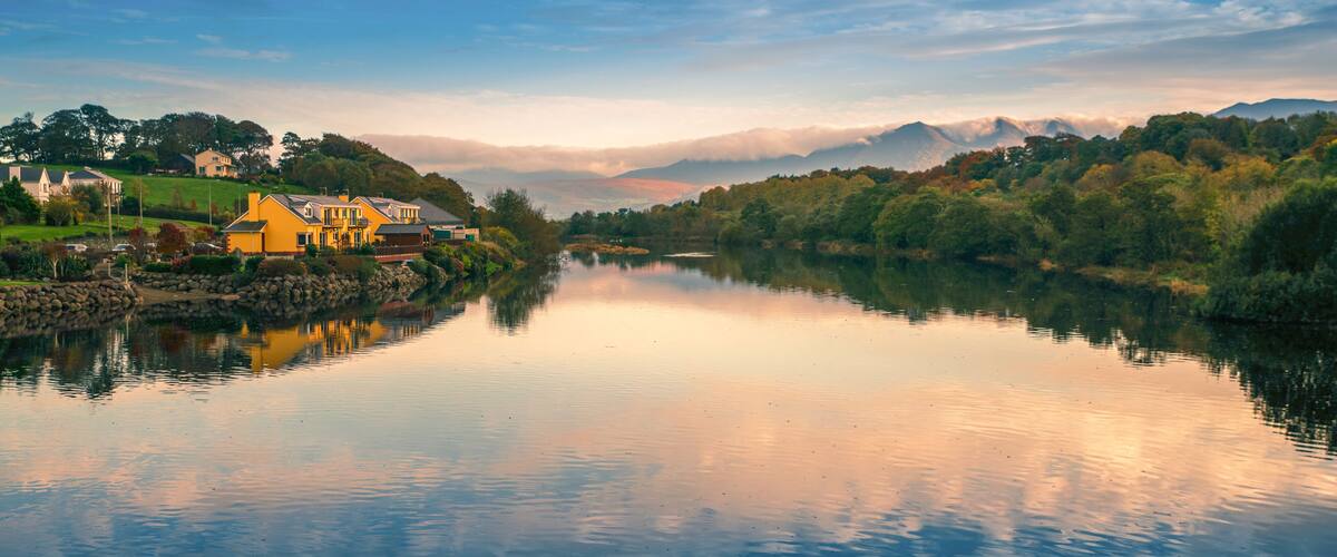 A calm river with a reflection of houses and trees, surrounded by mountains and a clear sky.