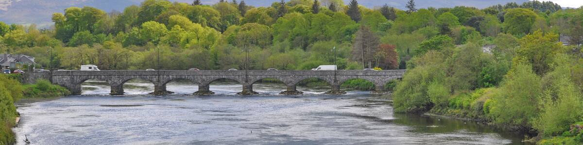 A river with a stone bridge, lush greenery, and mountains in the background.