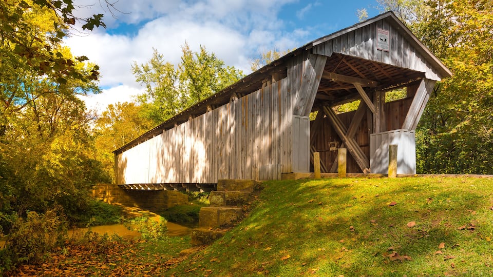 A wooden covered bridge in a wooded area with a grassy hill and a small stream.