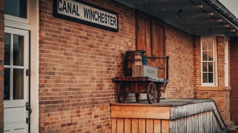 A brick building with a sign reading 'CANAL WINCHESTER' and a wooden cart with barrels.