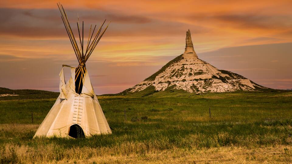 A teepee in a grassy field with a large hill in the background.
