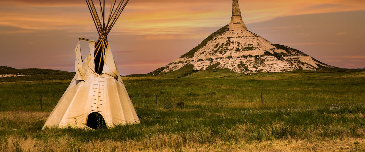 A teepee in a grassy field with a large hill in the background.