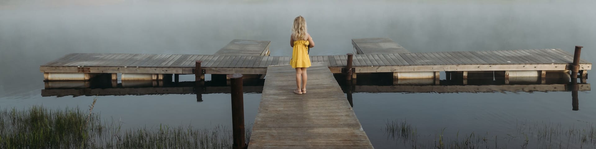 Girl in yellow dress stands on dock facing foggy lake in morning