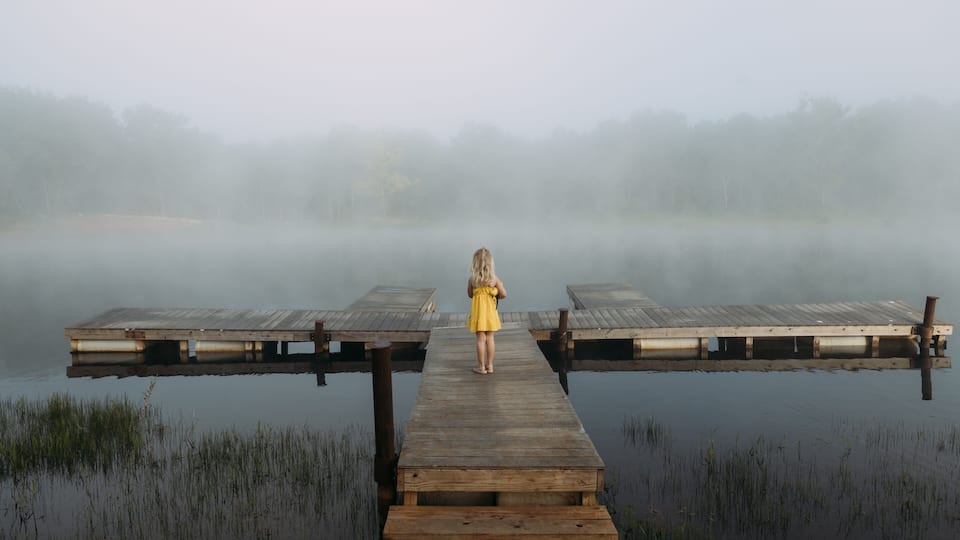 A wooden pier extending into a misty lake with a person in a yellow dress standing at the end.