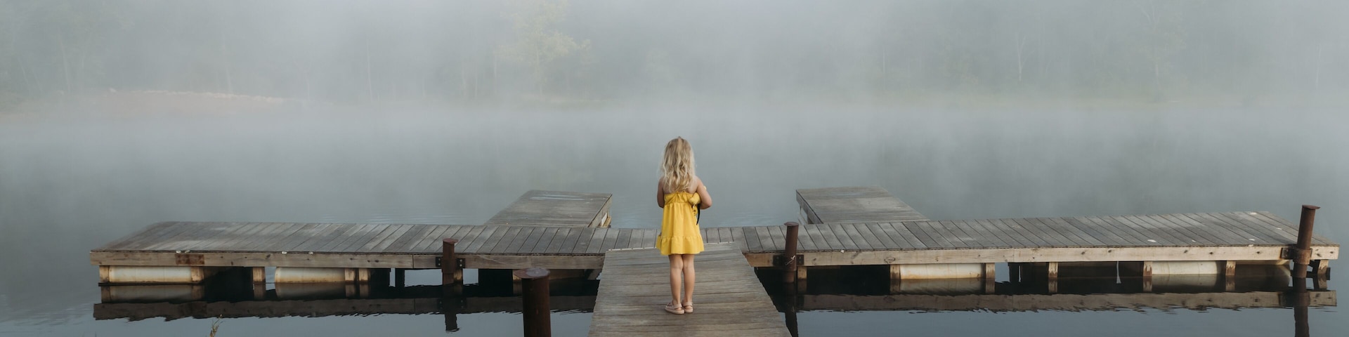 Girl in yellow dress stands on dock facing foggy lake in morning