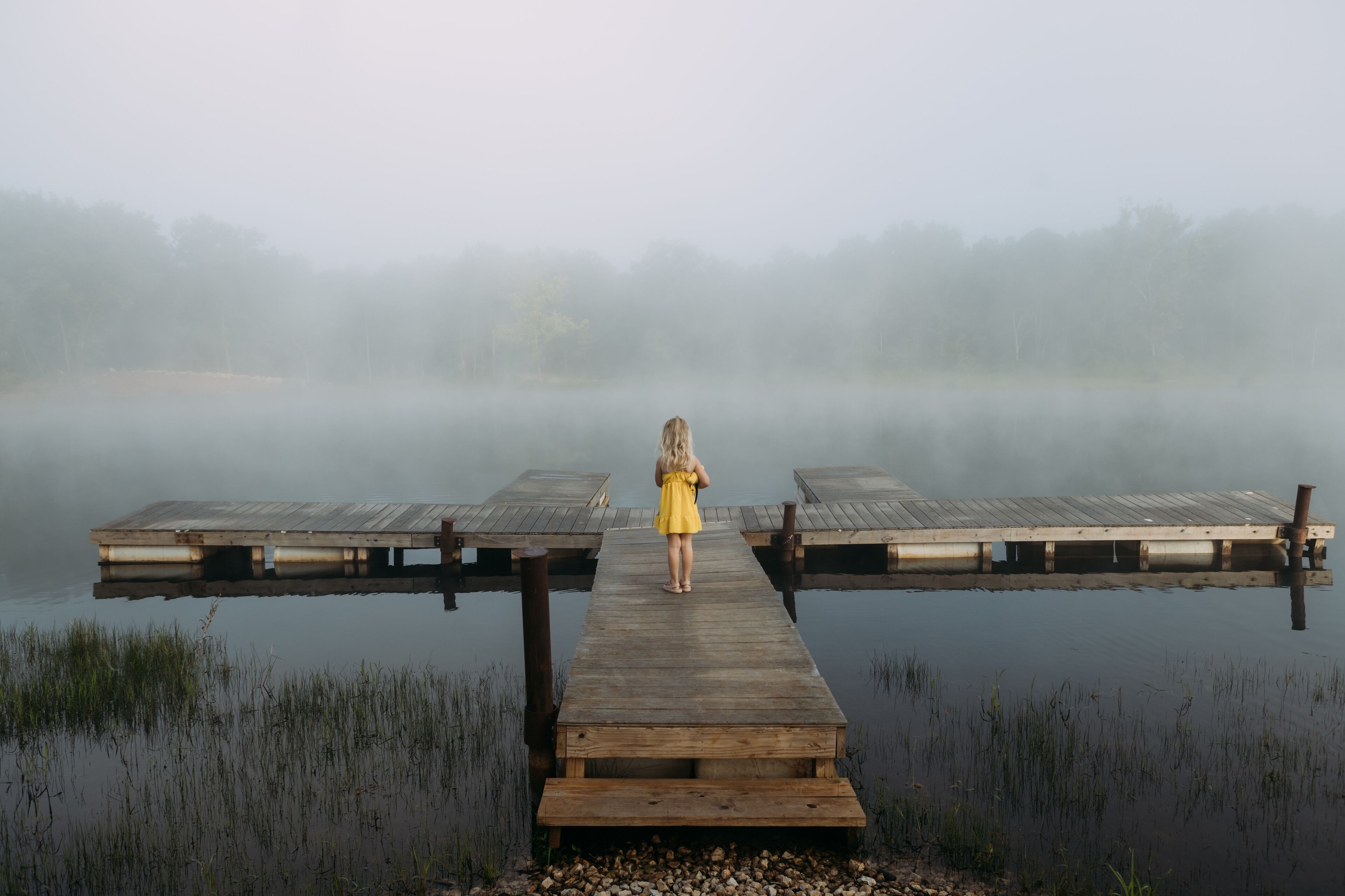 A wooden pier extending into a misty lake with a person in a yellow dress standing at the end.