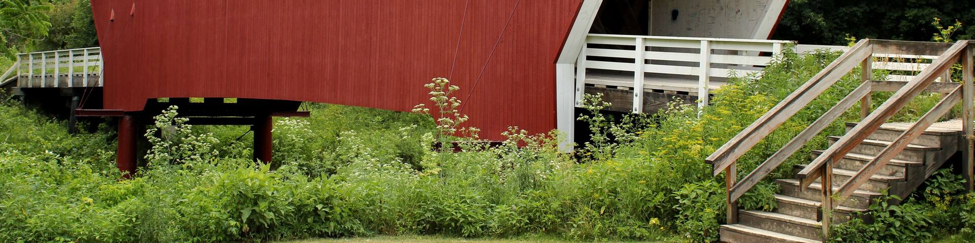 A red covered bridge with a white entrance, surrounded by greenery and a wooden staircase.