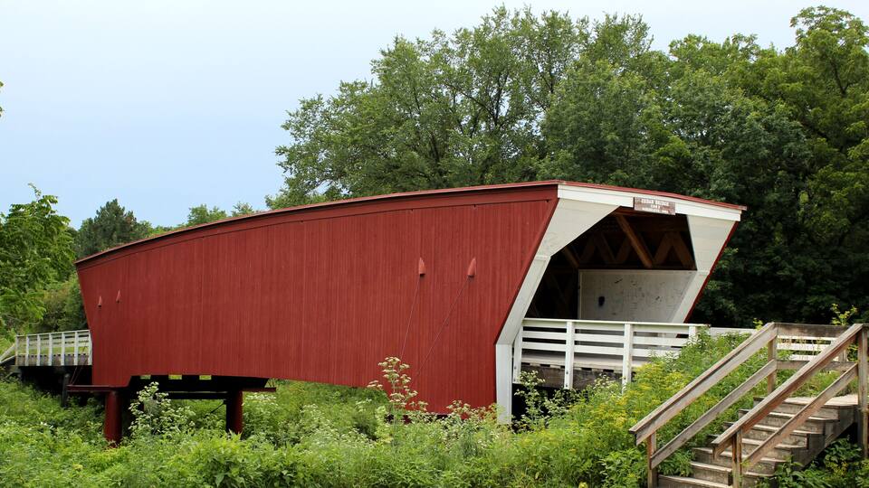 A red covered bridge with a white entrance, surrounded by greenery and a wooden staircase.