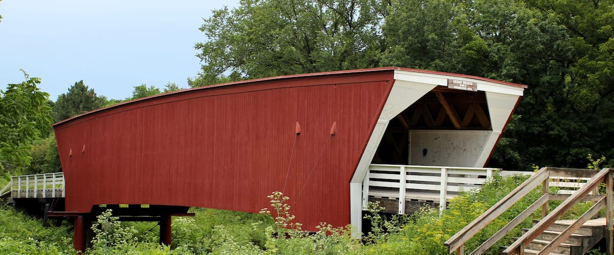 A red covered bridge with a white entrance, surrounded by greenery and a wooden staircase.