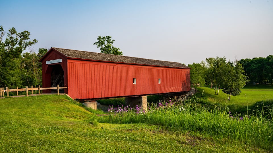 A red covered bridge with a wooden fence in a grassy field with trees in the background.