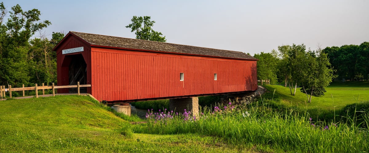A red covered bridge with a wooden fence in a grassy field with trees in the background.