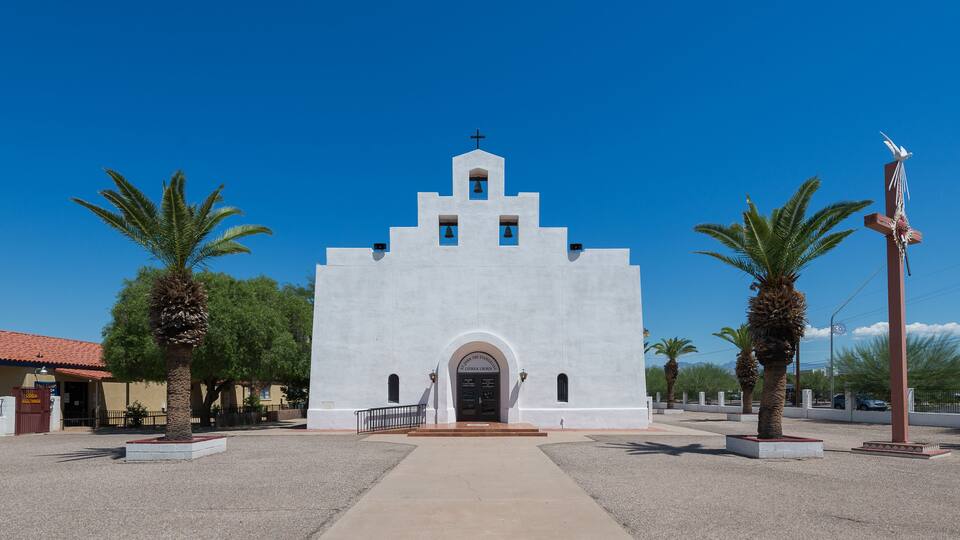 A white church with a bell tower, palm trees, and a cross sculpture.