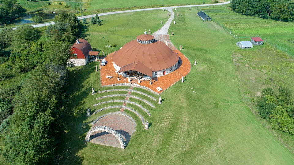 An aerial view of a large, circular building with a red roof, surrounded by a landscaped area with stone steps leading to a central point.