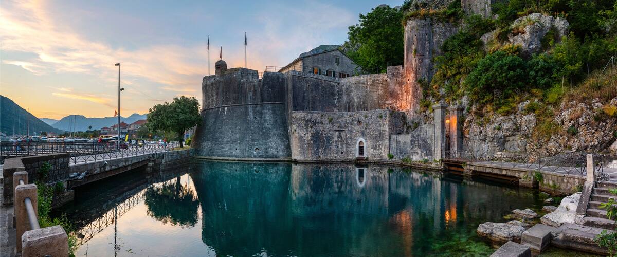 A historic stone fortress by a calm body of water, with a bridge leading to it and mountains in the background.
