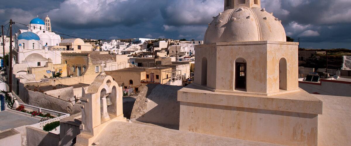 A coastal town with white buildings and blue domes under a cloudy sky.