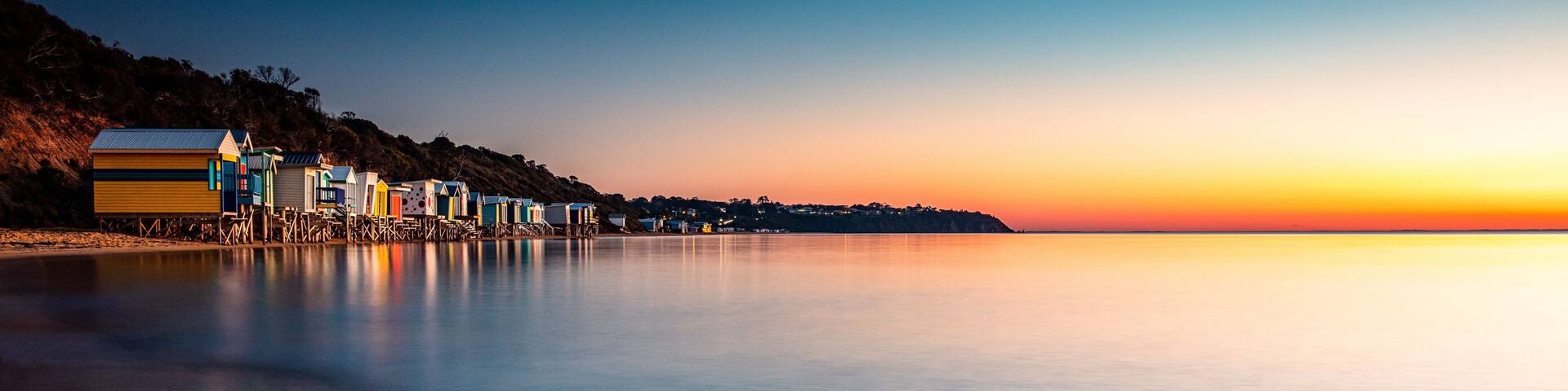 A coastal scene with colorful beach huts and a calm sea at sunset.