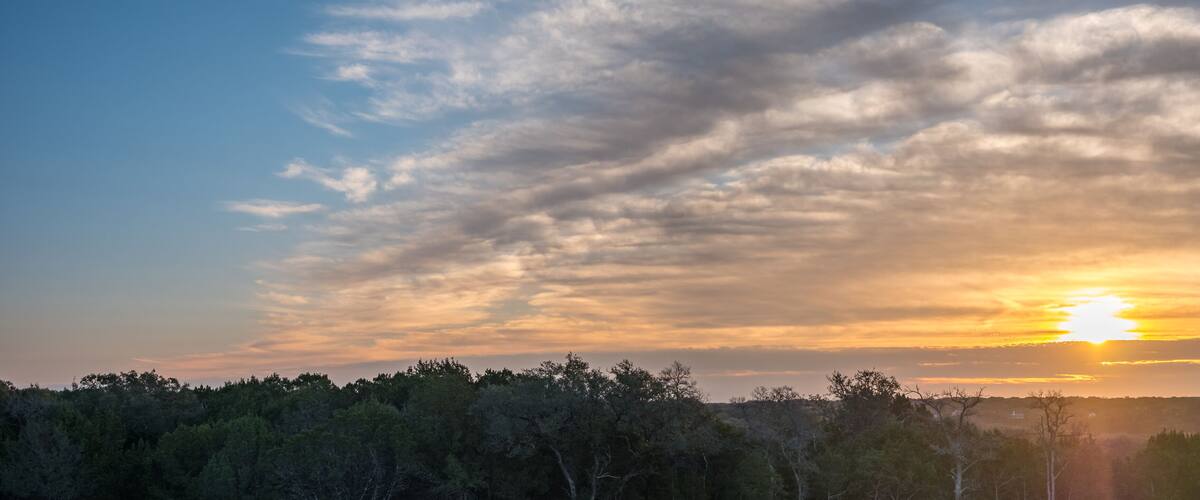 A sunset over a forested landscape with a clear sky and scattered clouds.
