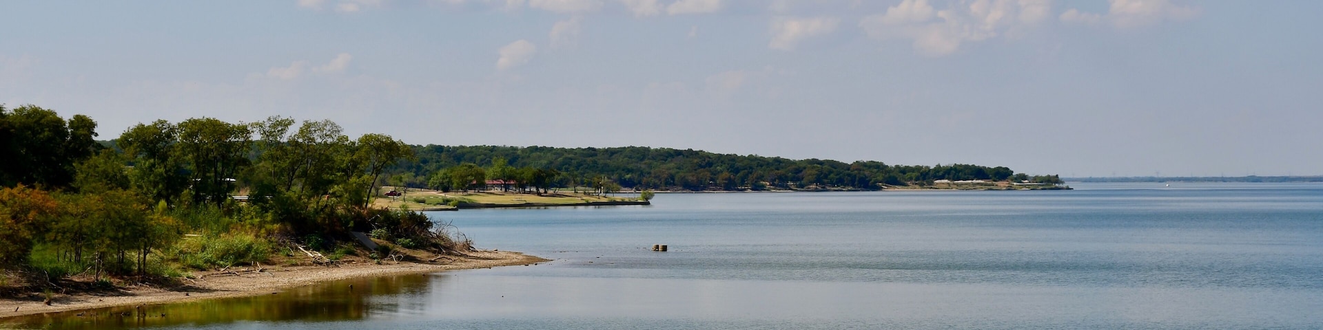 Un lago tranquilo con una orilla lejana, rodeado de árboles y un cielo despejado.