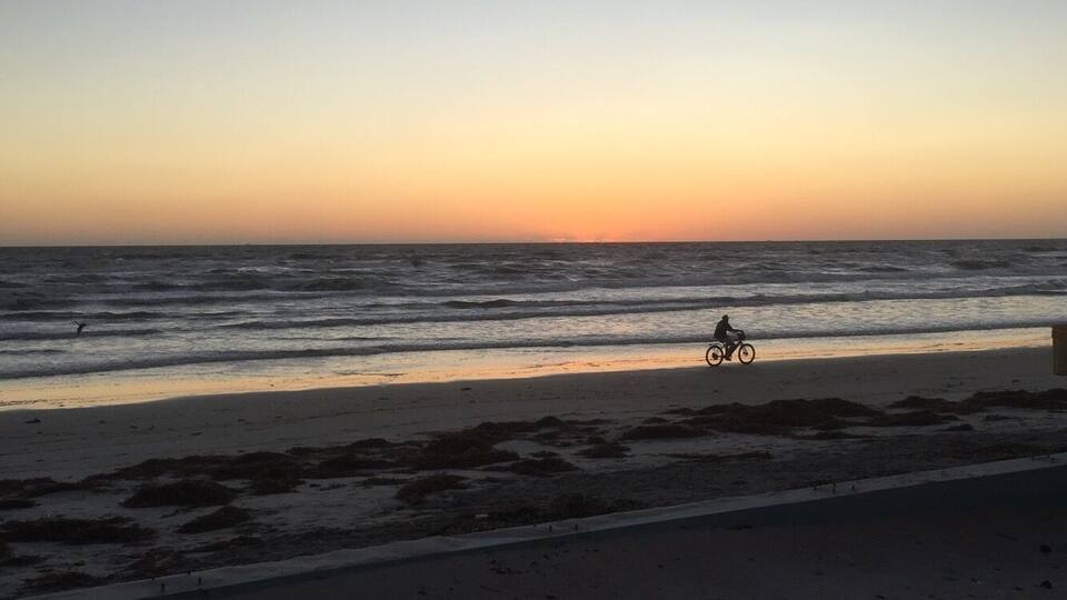 Ein Strand bei Sonnenuntergang mit Wellen, eine Person auf einem Fahrrad und ein ferner Sonnenuntergang.