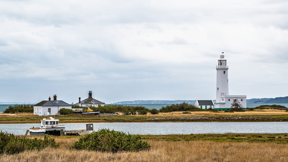 A coastal landscape with a lighthouse, a small white building, and a boat docked near the shore.