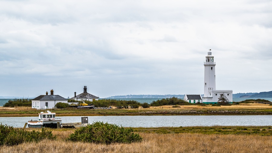 Un paisaje costero con un faro, un pequeño edificio blanco y un barco amarrado cerca de la orilla.