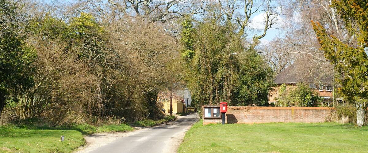 A rural road with a mailbox on the right, surrounded by trees and a brick wall.