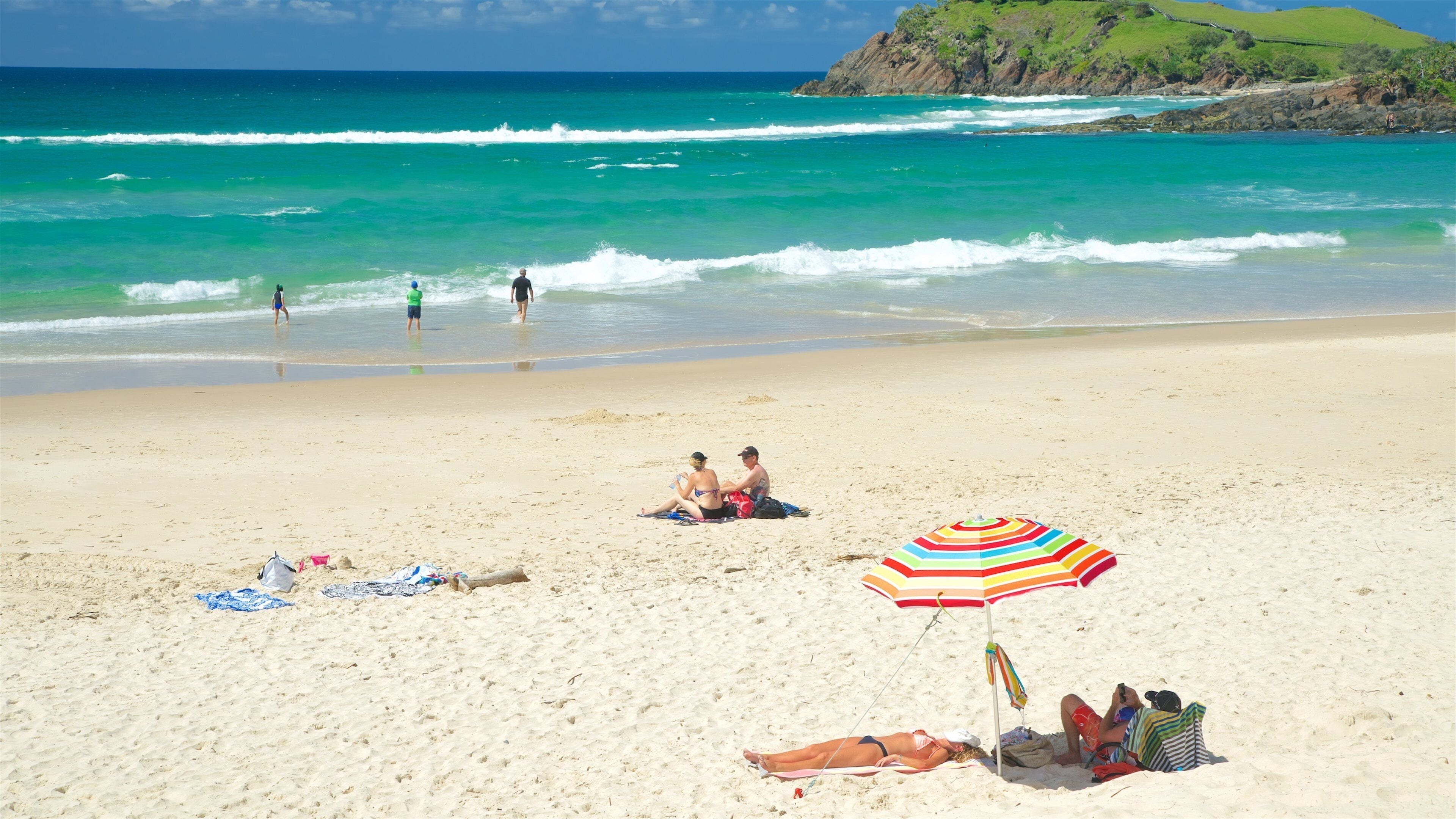 A beach with clear turquoise water, people sunbathing, and a colorful umbrella.