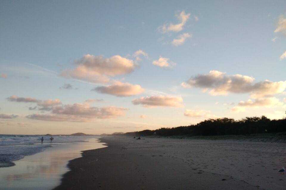A beach with a calm sea, a few people walking, and a clear sky with scattered clouds.