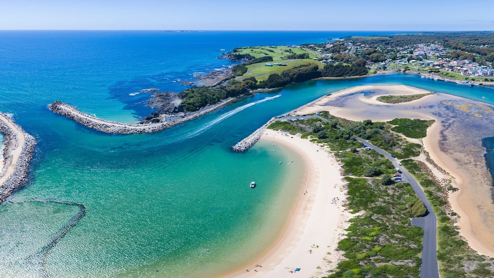 A coastal landscape with a curved road, a beach, and a rocky breakwater.