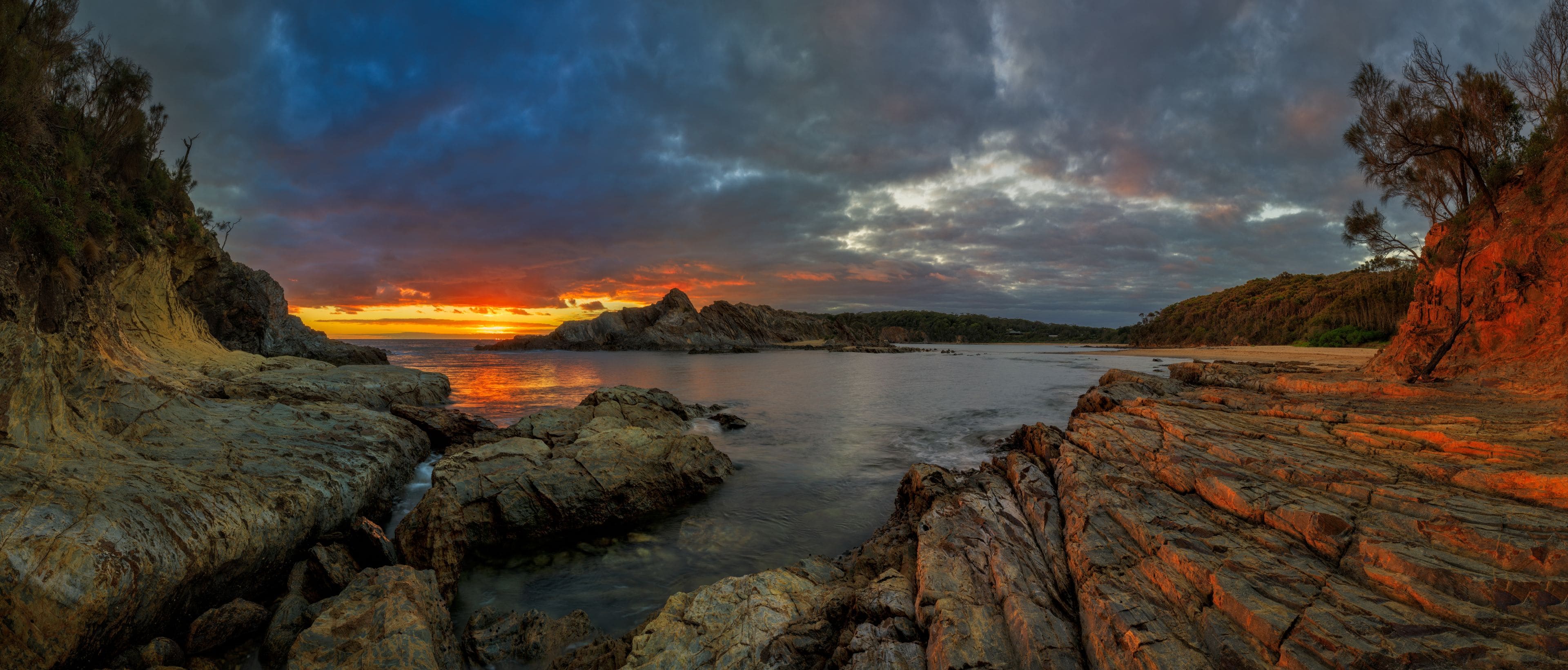 A coastal landscape with rocky terrain, a calm sea, and a dramatic sunset.