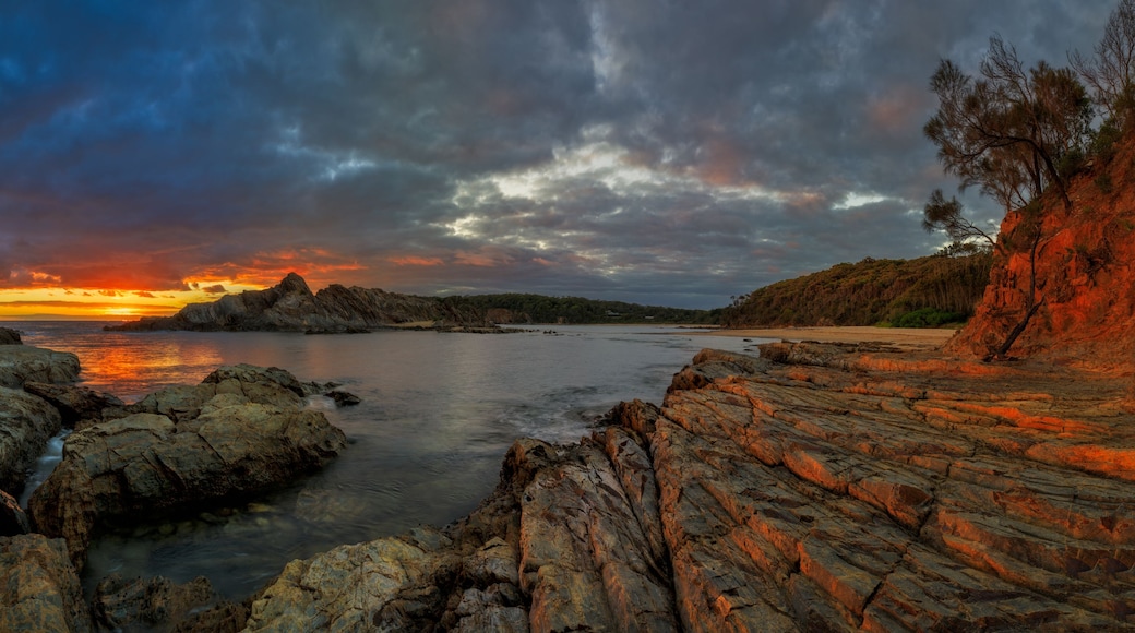 A coastal landscape with rocky terrain, a calm sea, and a dramatic sunset.