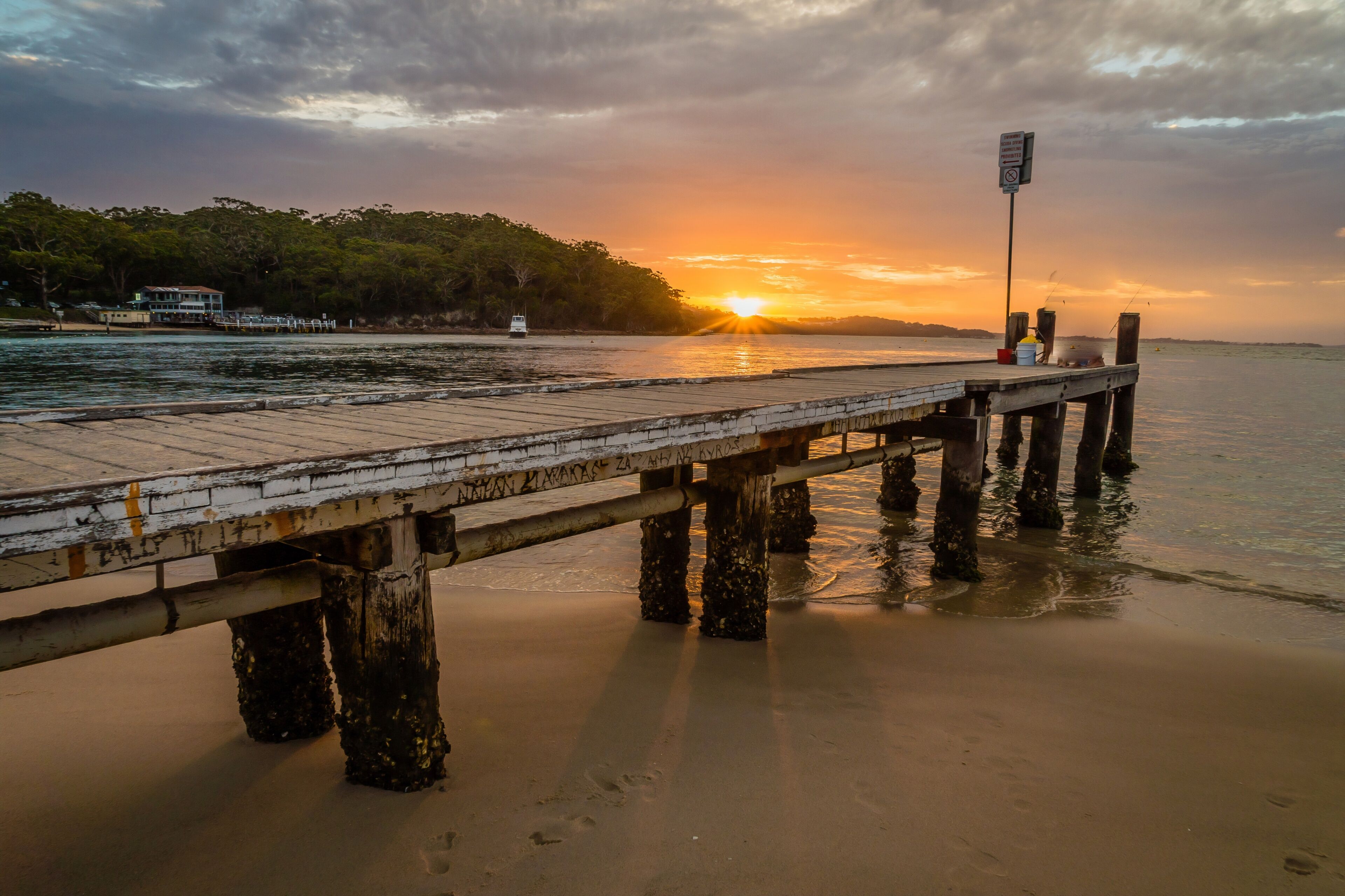 A wooden pier extending into calm waters at sunset, with a boat on the left and a signpost on the right.