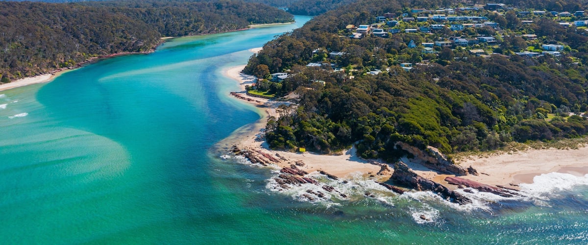 A coastal landscape with turquoise waters, a sandy beach, and a residential area surrounded by dense forest.