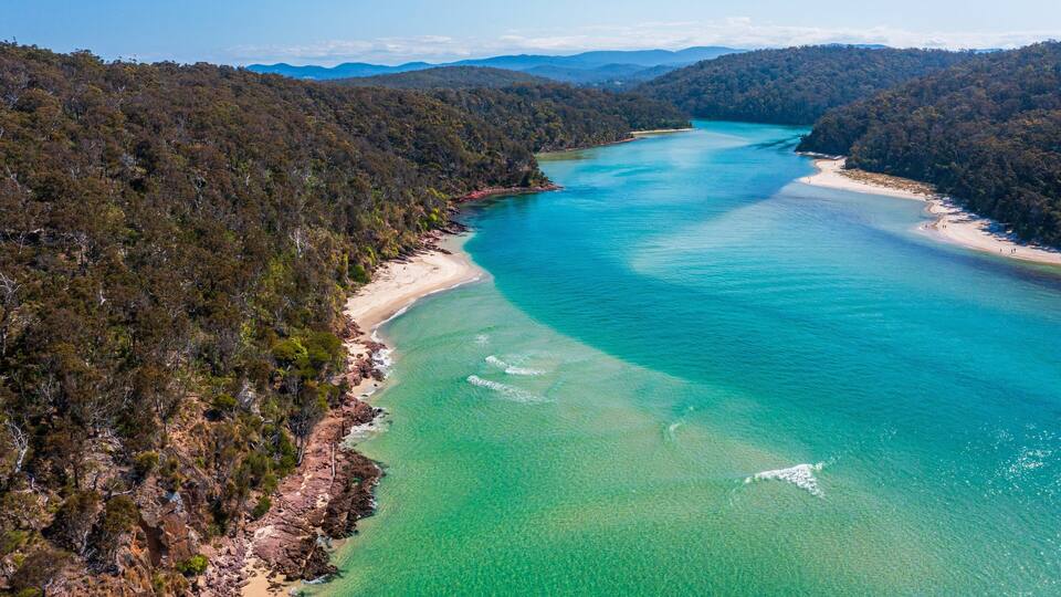 A coastal landscape with a clear turquoise sea, sandy beaches, and dense forested hills.