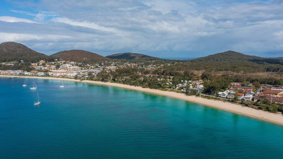 A coastal town with a beach, clear blue waters, and hills in the background.