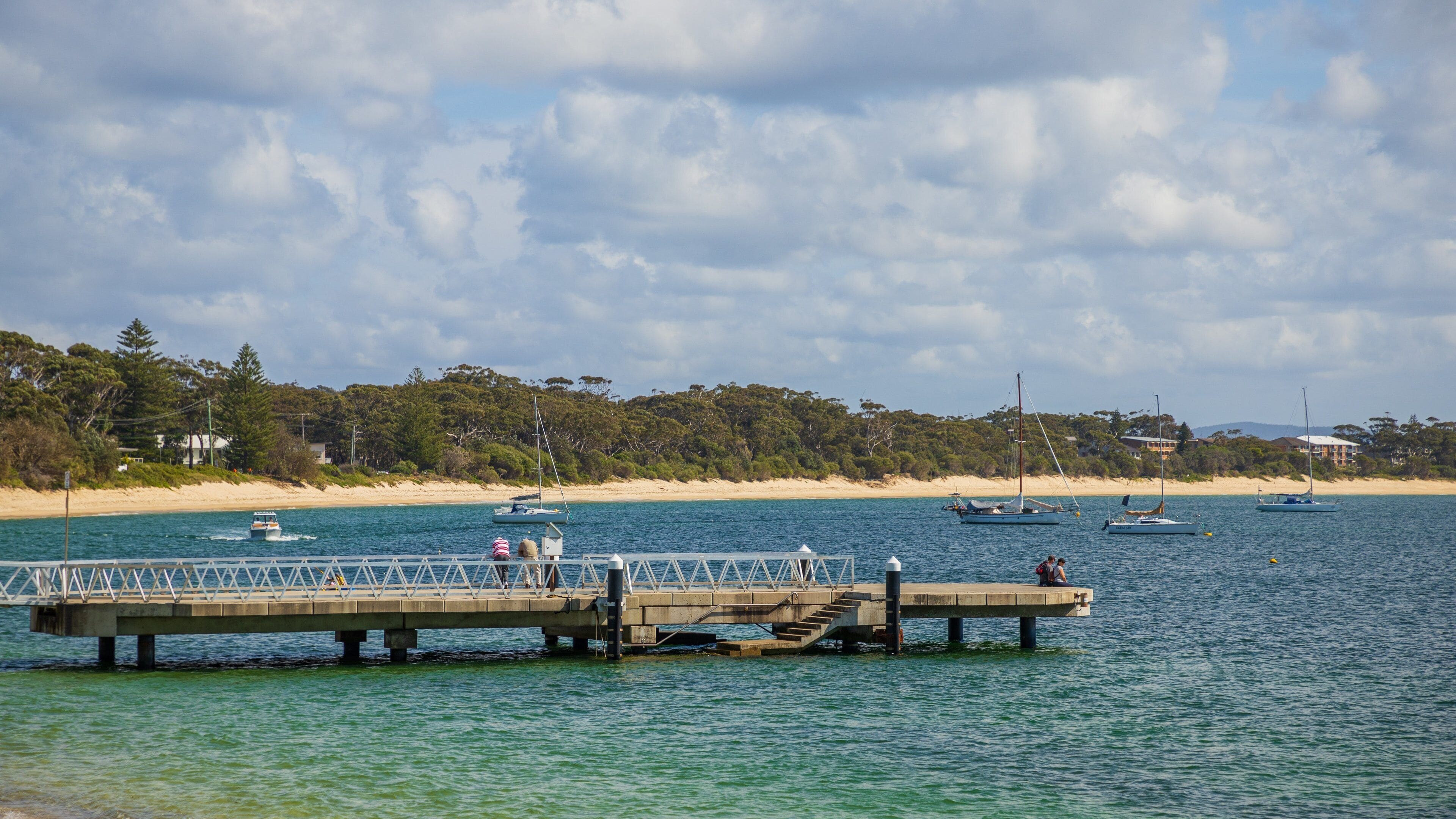 A pier extending into calm waters with boats and a beach in the background.