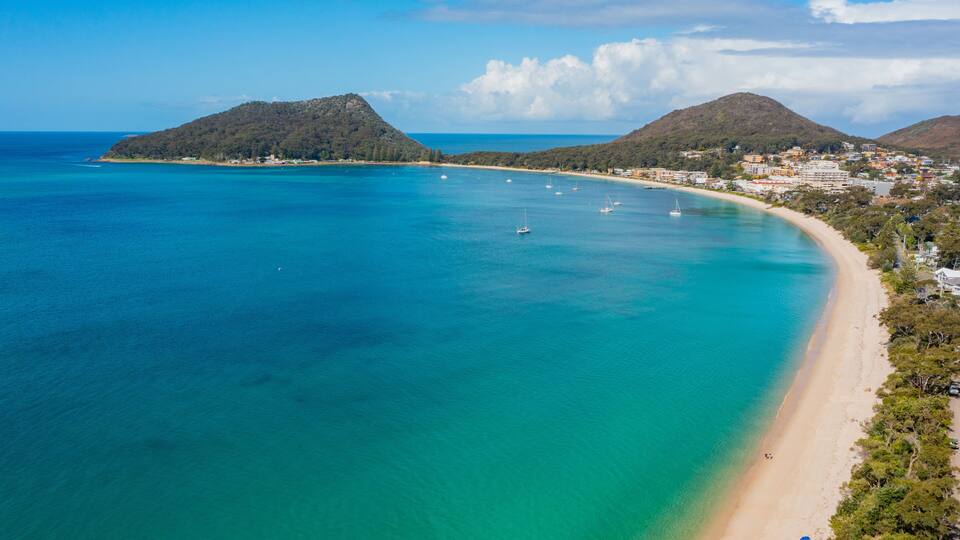 A coastal landscape with a sandy beach, clear turquoise waters, and two prominent hills in the background.