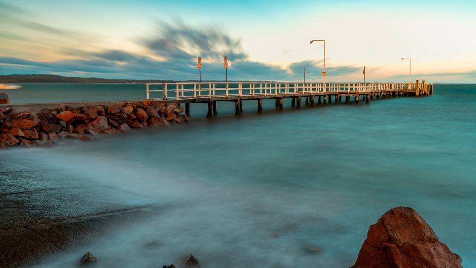 A pier extending into calm waters, surrounded by rocks and a cloudy sky.
