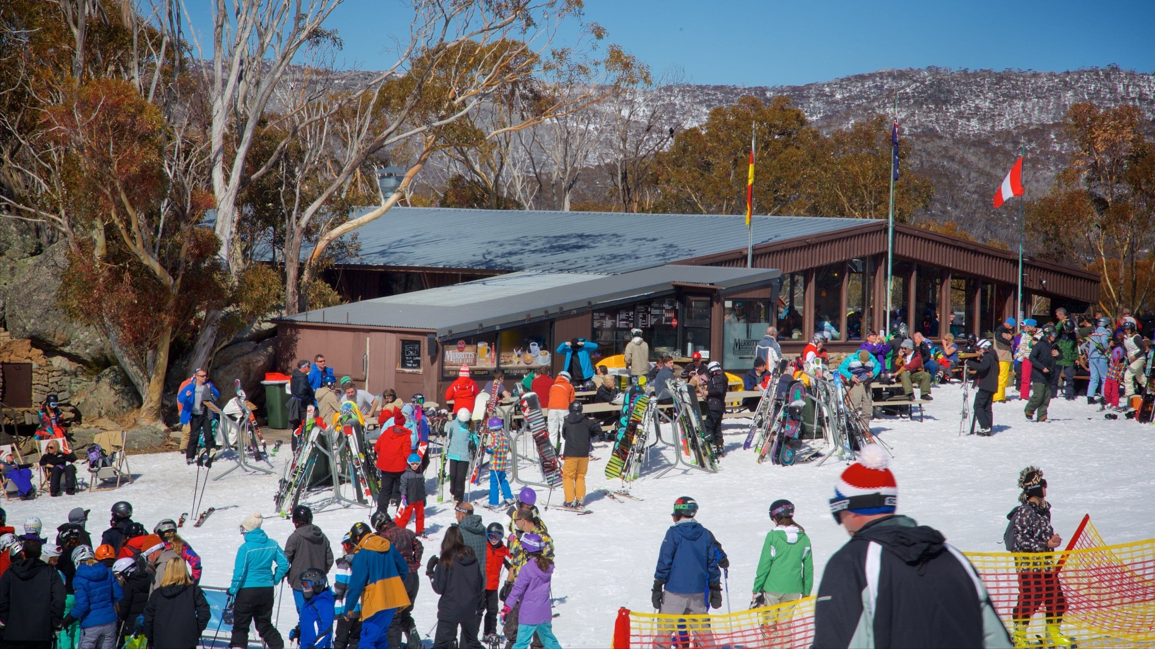 A snowy ski resort with a crowd of people, ski equipment, and a building with flags.
