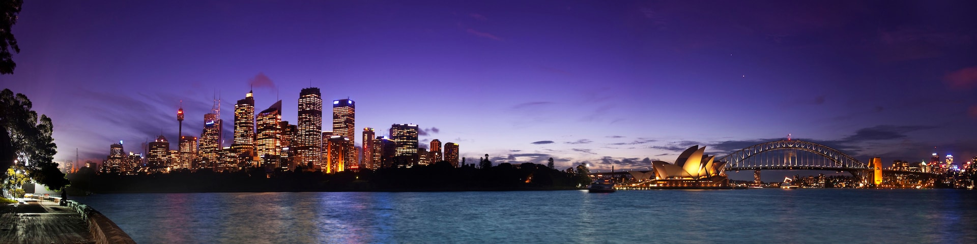Ein Stadtpanorama im Abendlicht mit beleuchteten Gebäuden und dem Sydney Opera House.