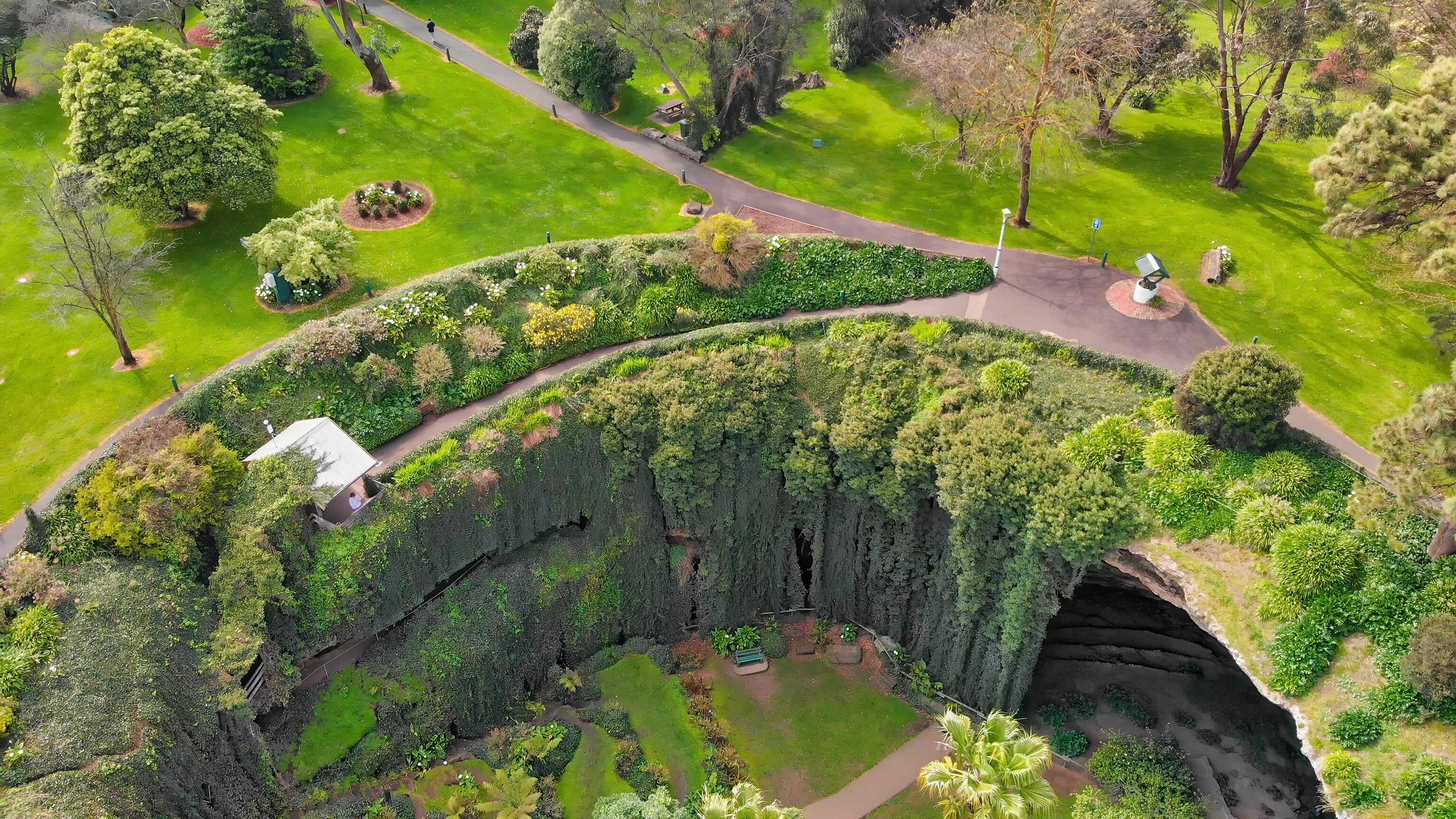 An aerial view of a park with a curved pathway, lush greenery, and a small white building.