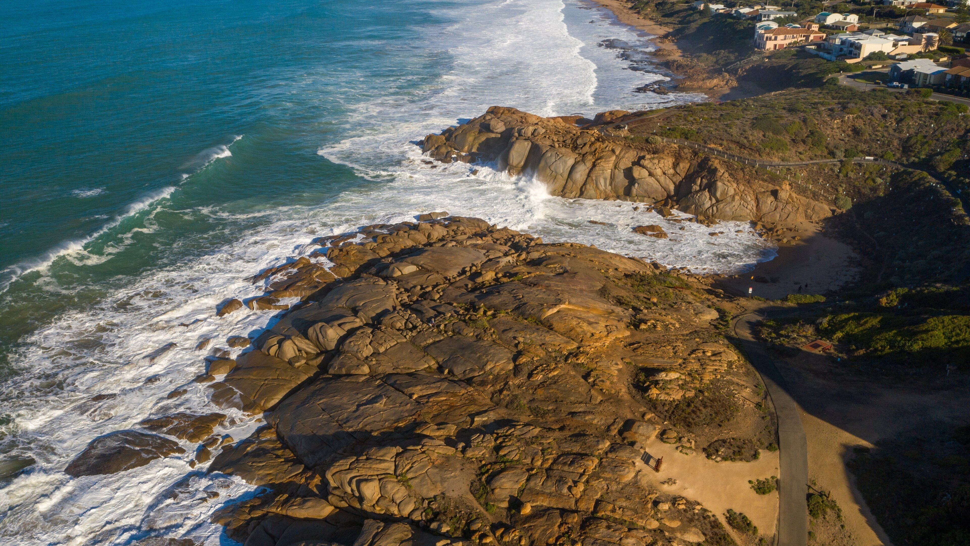 A coastal landscape with rocky cliffs, waves crashing against the shore, and a road leading to a residential area.